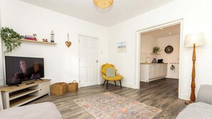 Uma sala de estar com televisão, uma cadeira amarela, um tapete estampado e uma cozinha ao fundo.
