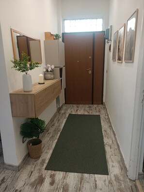 Een hal met een houten vloer, een groene mat, een houten kast, een spiegel, een kamerplant, een deur en lijstjes met foto's aan de muur.