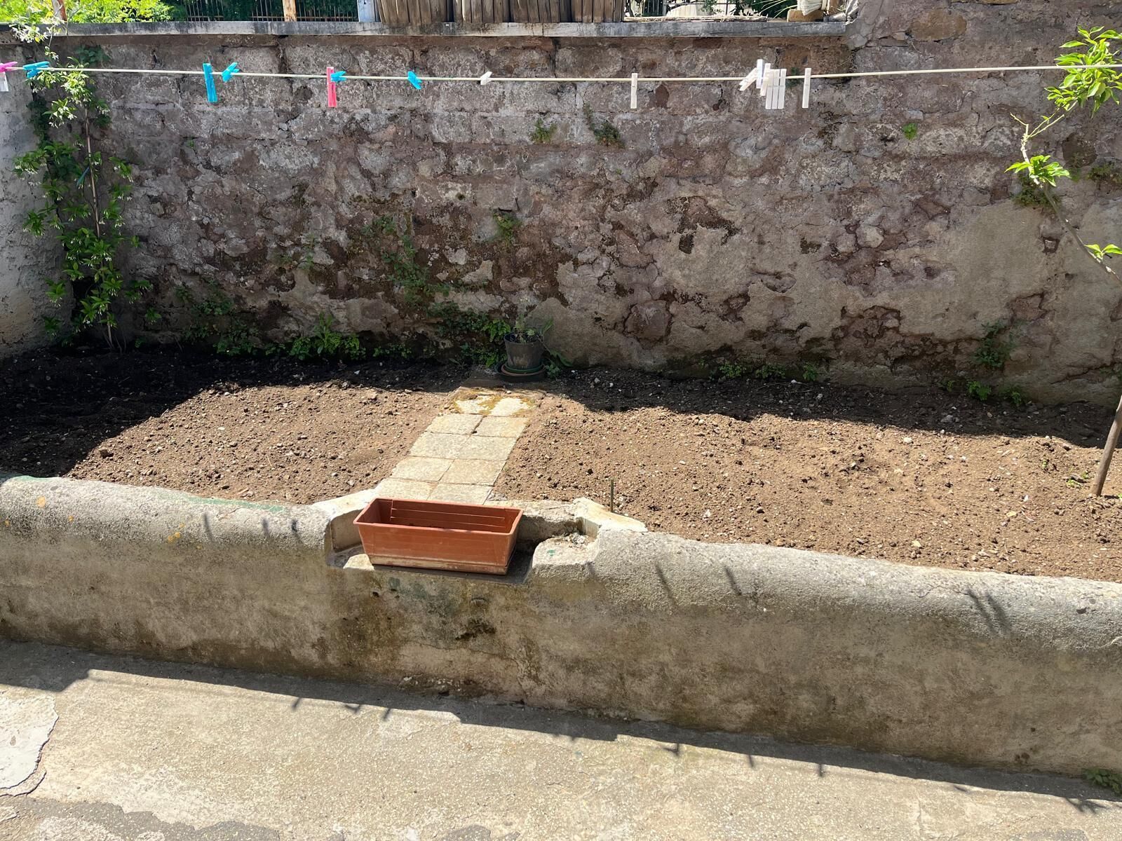 A garden with a concrete border, a wooden planter, and a clothesline with clothespins against a stone wall.