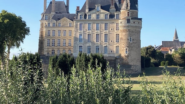 Ein großes, historisches Gebäude mit mehreren Türmen, darunter ein markanter zentraler Turm, umgeben von viel Grün.