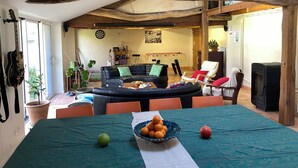 Ein geräumiger Wohnbereich mit hoher Decke, einem Esstisch mit Früchten, einer Couch und einem Kamin.