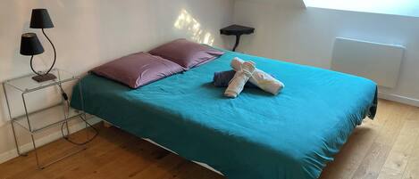 A bedroom with a teal bedspread, two pillows, a rolled towel, a lamp, a small side table, a radiator, and a skylight.