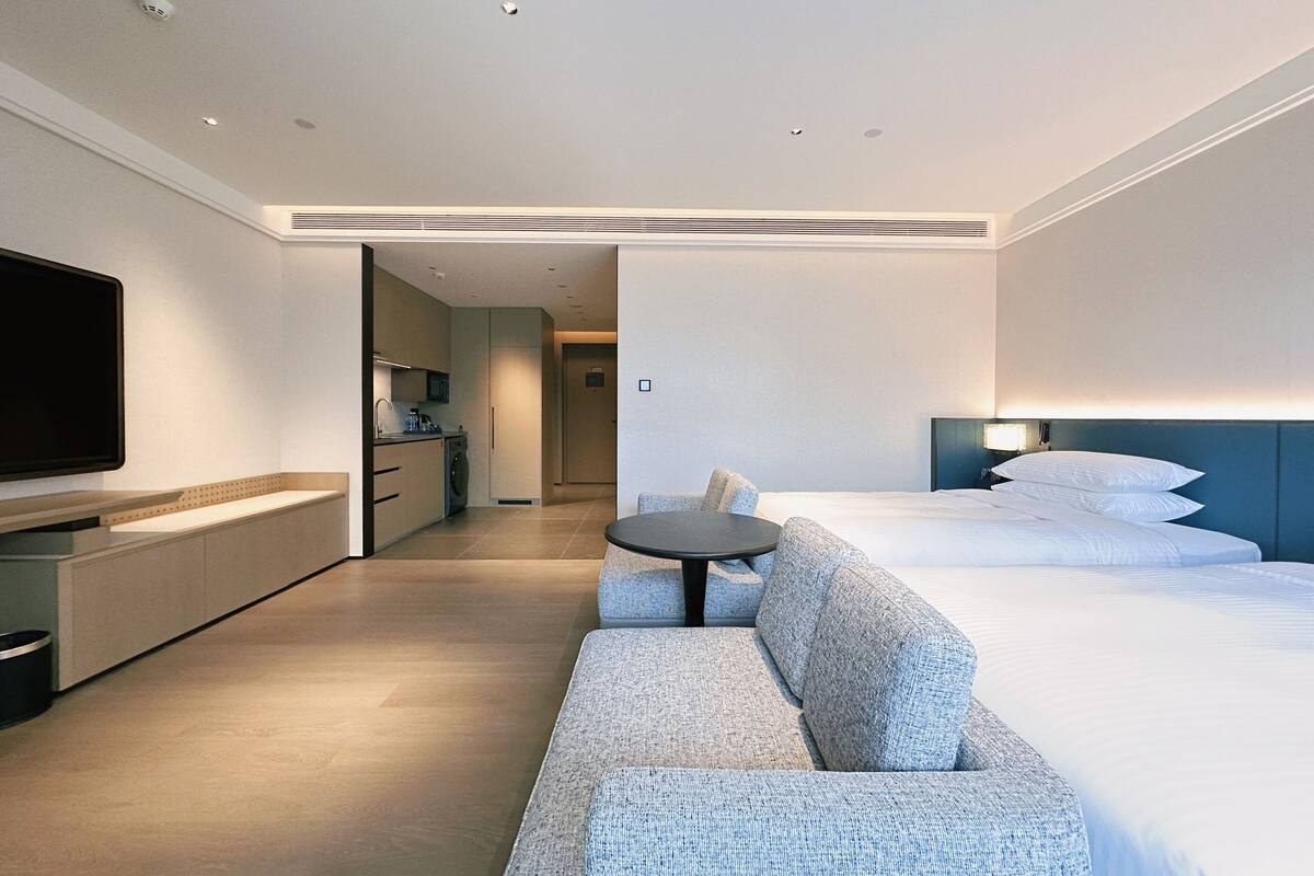 A modern hotel room with a sofa, a bed, a TV, a small table, and a kitchenette.