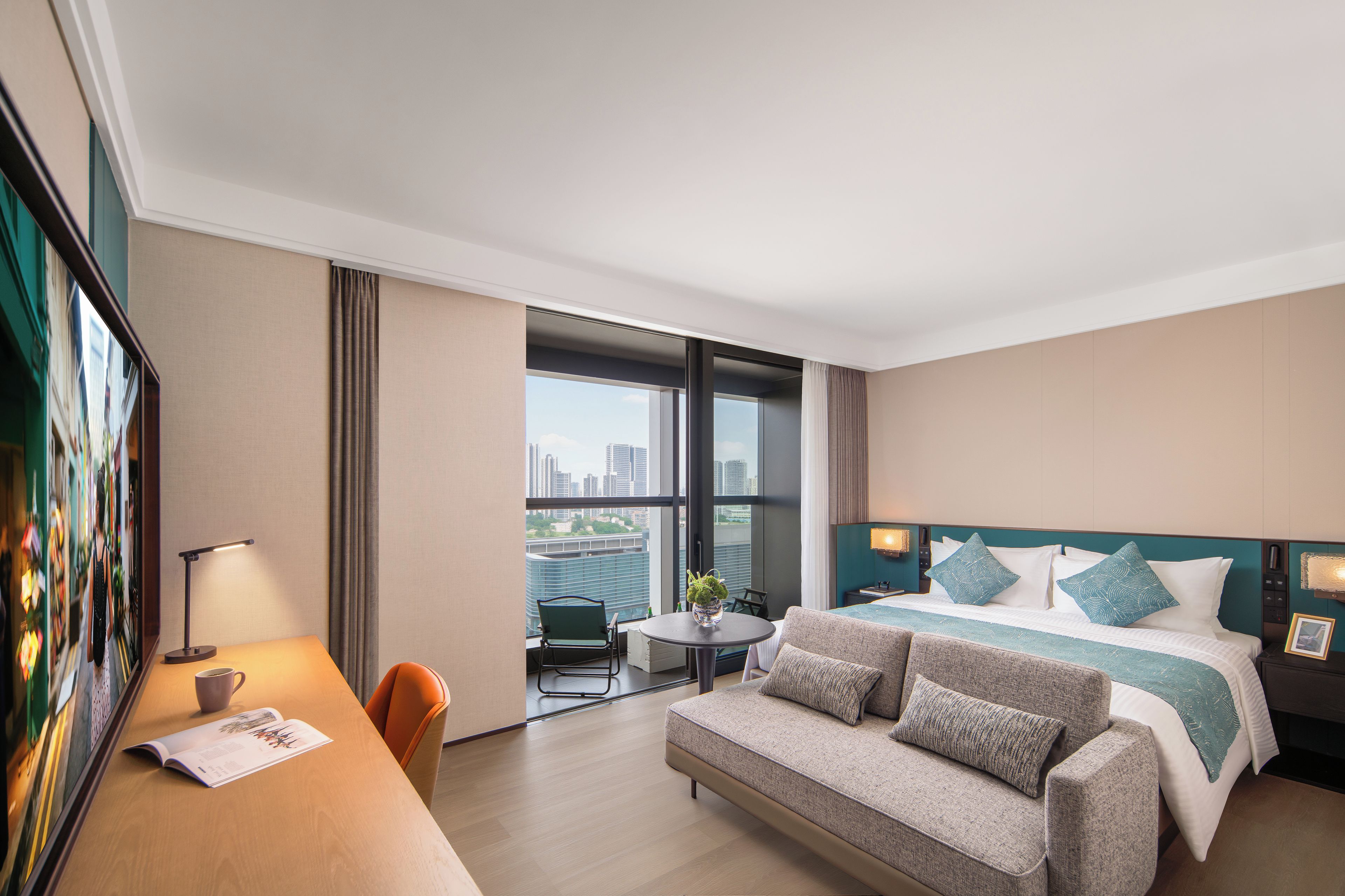 Een hotelkamer met een bed, een bank, een bureau en uitzicht op de stad door grote ramen.