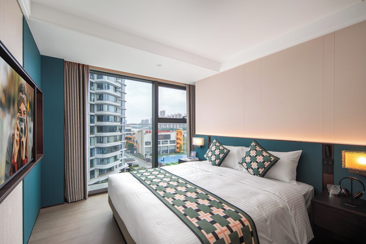 Habitación de hotel con una cama grande, una mesita de noche con lámpara, vista a un paisaje urbano a través de un ventanal amplio y un cuadro en la pared.
