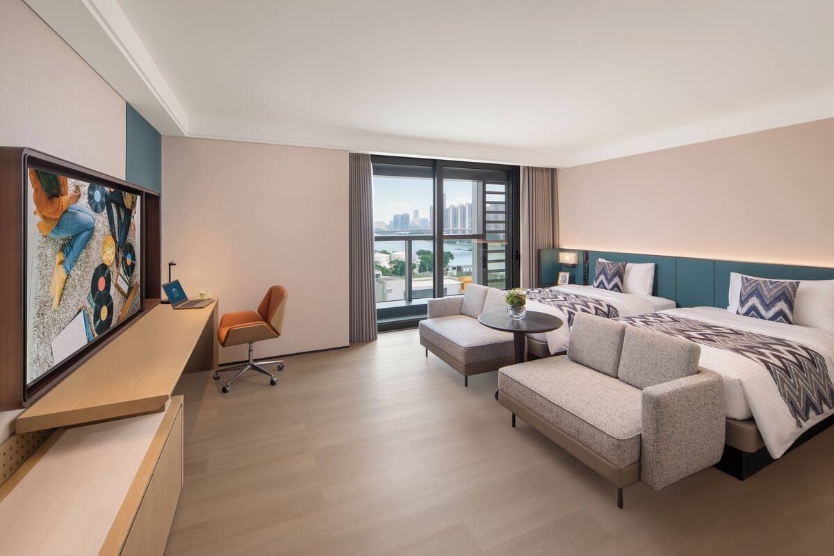 A modern hotel room with a bed, sofa, desk, and a large window with a city view.