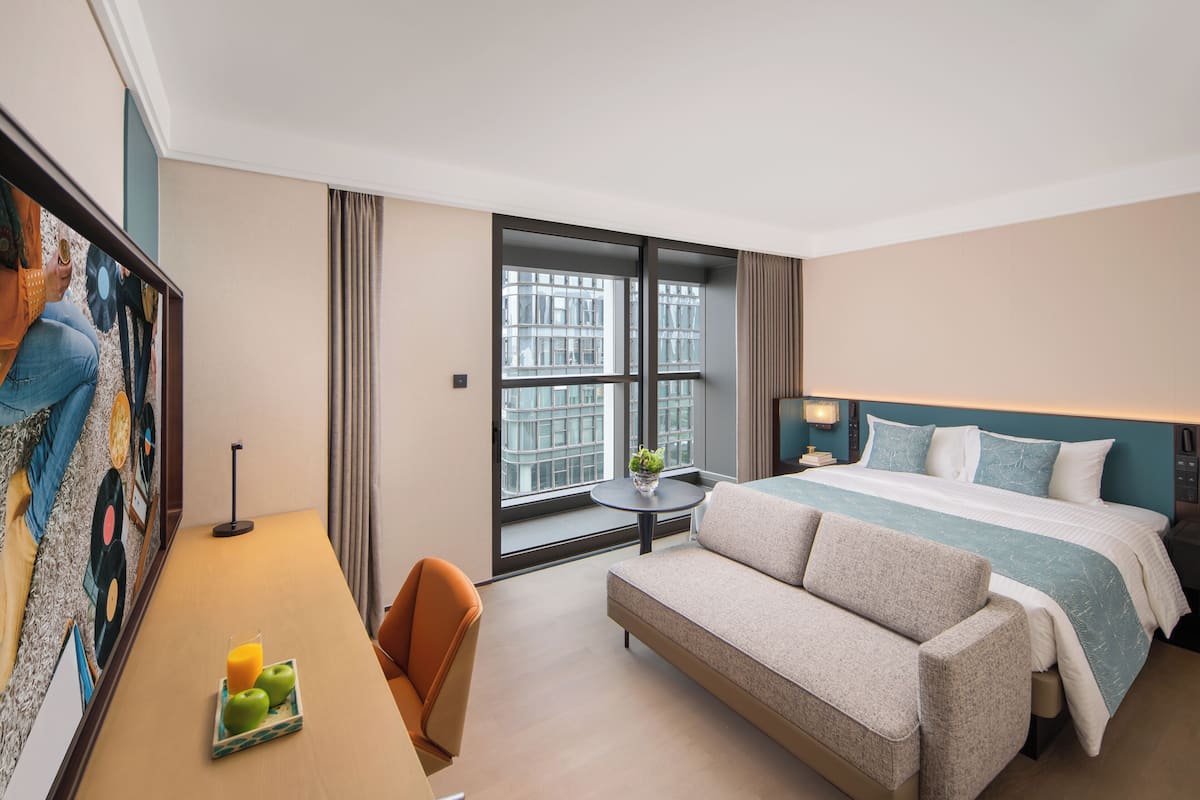 A hotel room with a bed, sofa, desk, and a large window with a city view.