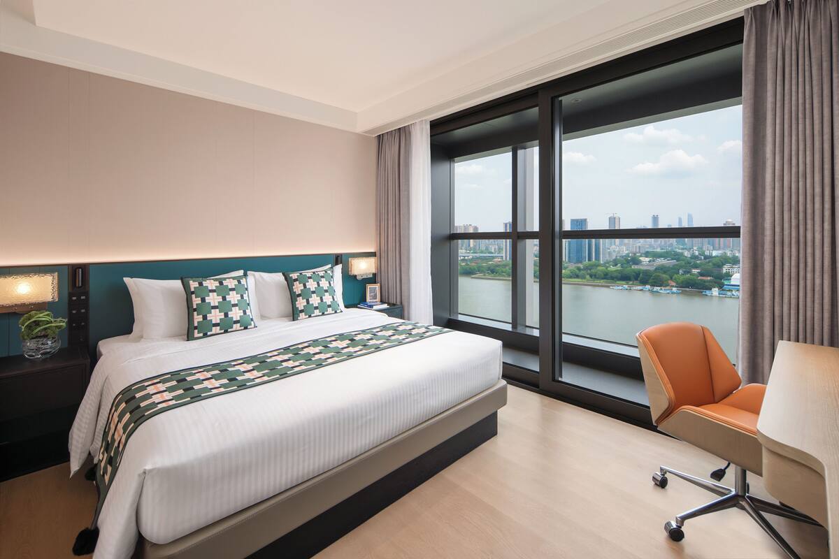 A hotel room with a large bed, a desk, a chair, a nightstand, and a view of a cityscape through large windows.