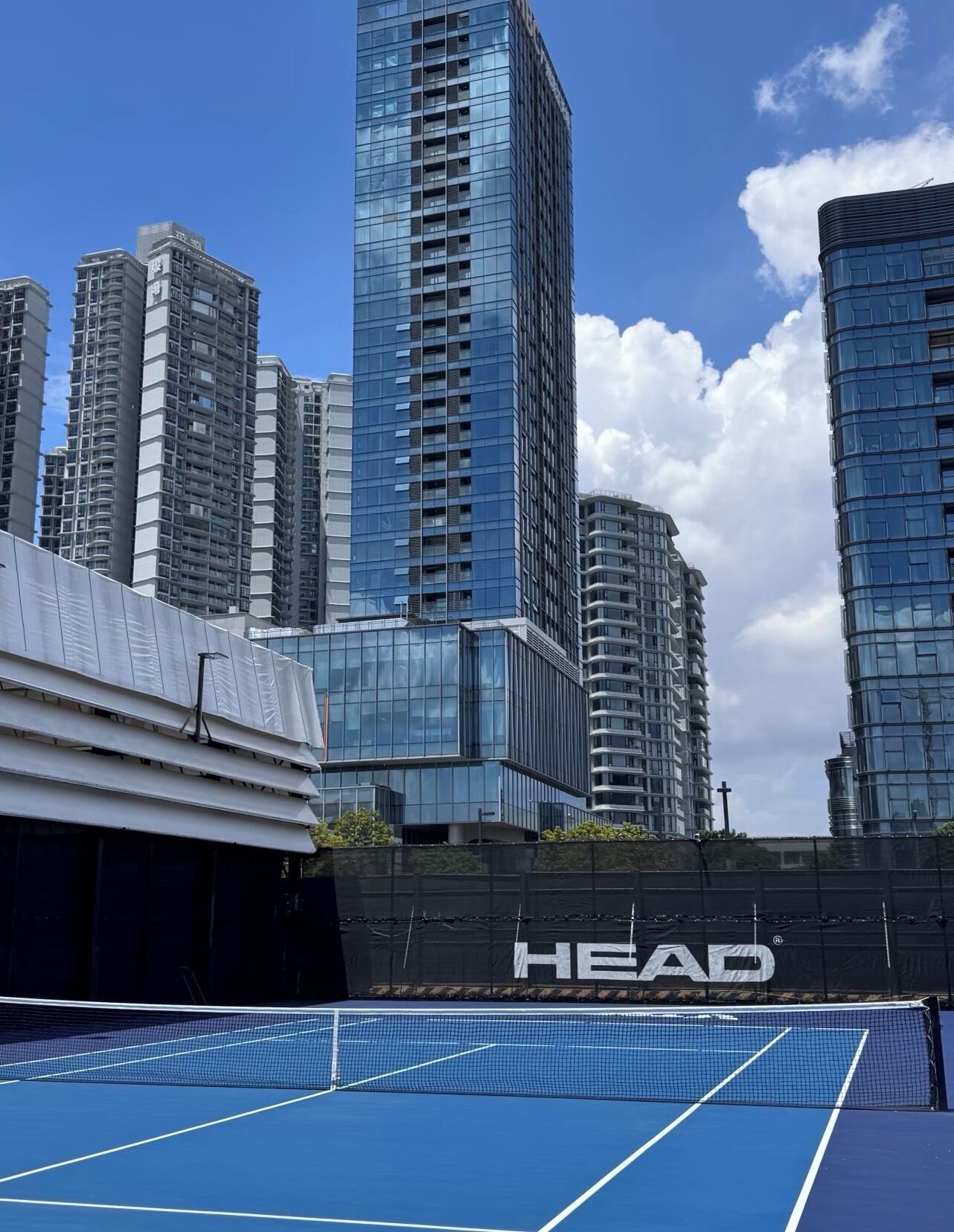 Una cancha de tenis con una red, rodeada de edificios altos y un muro negro con la palabra 'HEAD'.