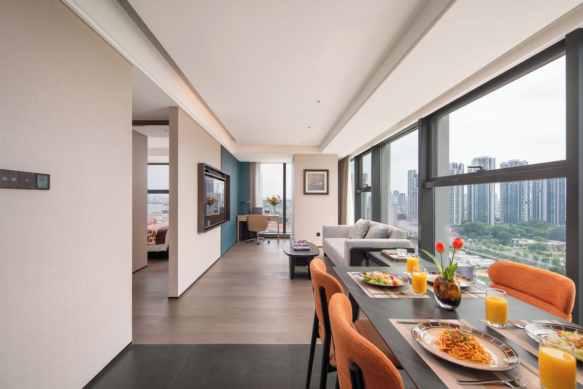 A modern hotel room with a dining area, large windows with a city view, and a living area with a sofa and table.