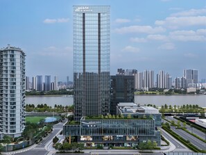 A modern high-rise building with a glass facade, surrounded by other high-rise buildings and a park area with a road and greenery.