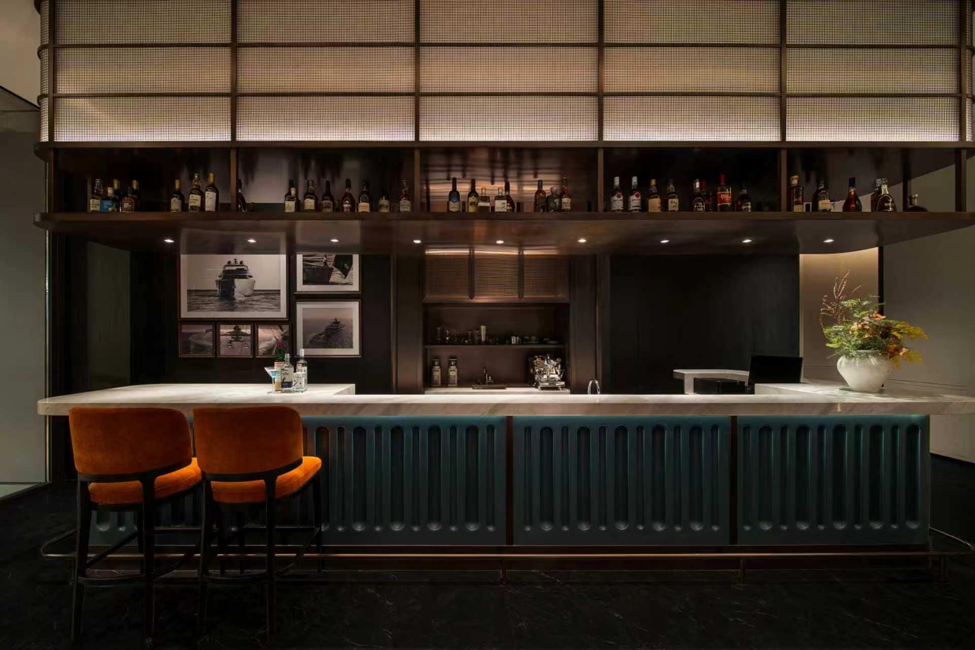 Un espace bar avec un comptoir, deux chaises orange, une plante en pot et des tableaux encadrés au mur.