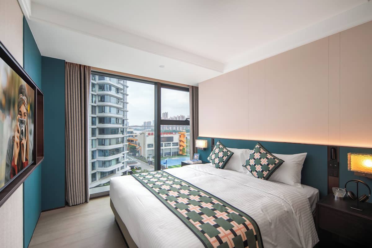 A hotel room with a large bed, a nightstand with a lamp, a view of a cityscape through a large window, and a painting on the wall.