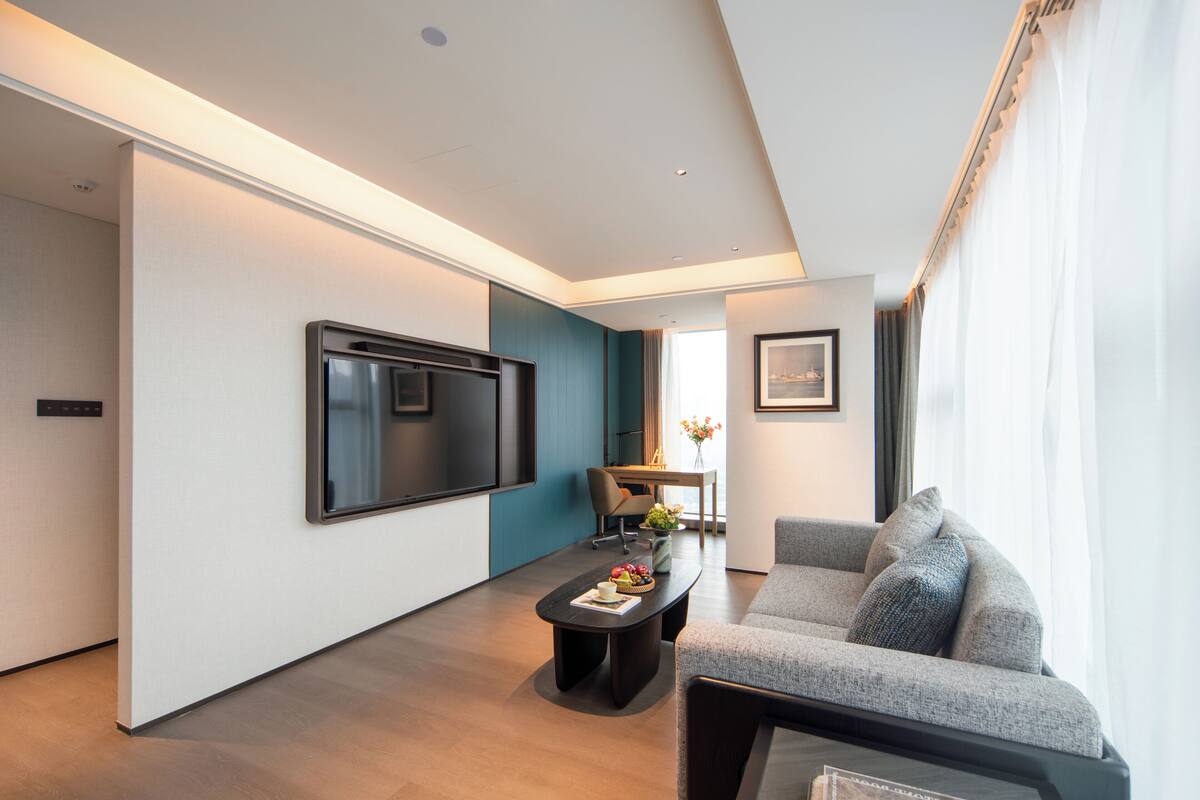 A modern living room with a flat-screen TV, a sofa, a coffee table with fruits, and a dining area with a table and chairs.