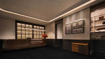 Un hall d'hôtel moderne comprenant un comptoir d'accueil, une grande fenêtre avec des stores verticaux, un écran fixé au mur et une vieille malle.