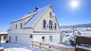 Une maison blanche à deux étages, avec un toit en pente, une clôture en bois et un ciel d’un bleu limpide.