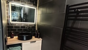 Um banheiro compacto com revestimento de azulejos pretos, um espelho, uma pia, porta-toalhas e uma pequena lixeira.