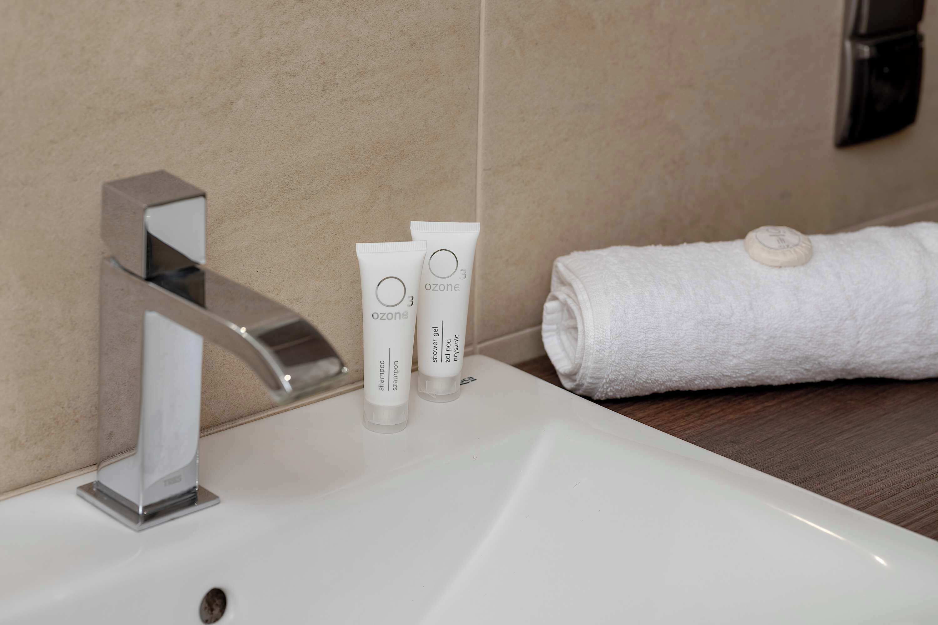 Une salle de bain avec un lavabo blanc, un robinet chromé, deux tubes de gel douche de la marque Ozone et une serviette blanche posée sur une surface en bois.