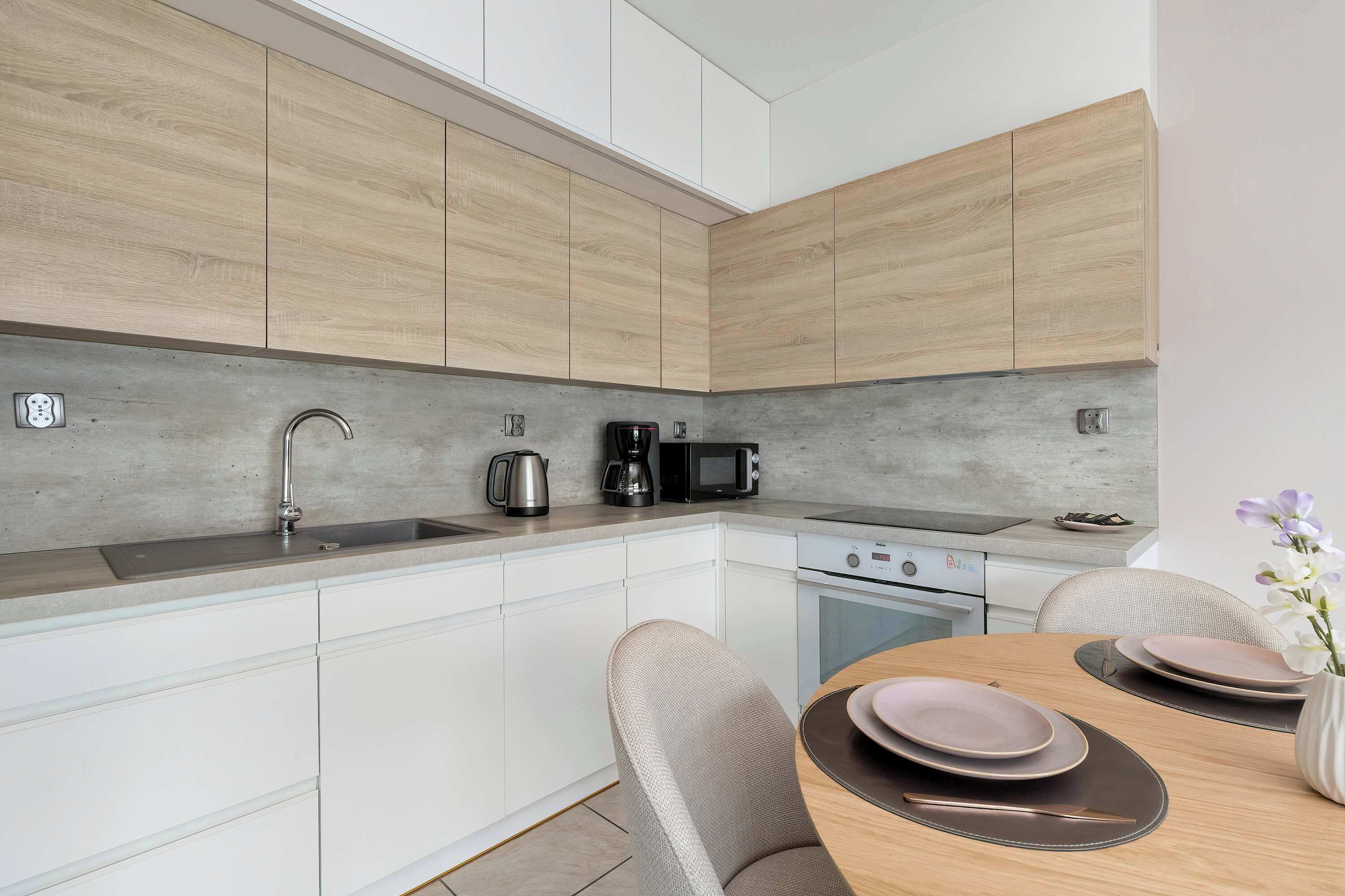Una cucina moderna con mobili bianchi, un piano di lavoro grigio, un lavello, un bollitore, una macchina del caffè, un forno a microonde e una stufa. C'è un tavolo da pranzo con piatti e un vaso con fiori.