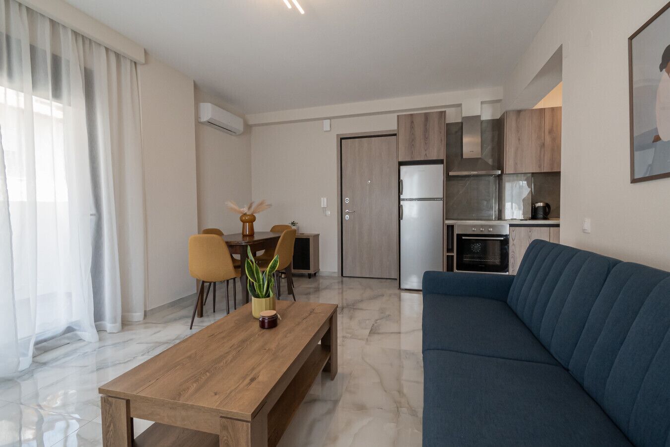 Een moderne woonkamer met een houten salontafel, een blauwe bank, een eethoek met stoelen, en een keuken met een koelkast en oven.
