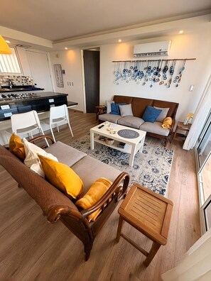 A modern living room with a sofa, coffee table, and chairs, a kitchen area with a dining table, and a decorative wall piece.