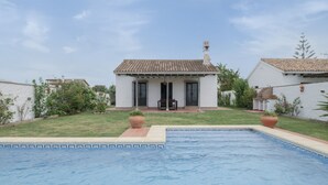 Ein Pool mit einer gefliesten Kante, ein weißes Gebäude mit einem geflasten Dach und eine kleine Terrasse mit Stuhl und Tisch.