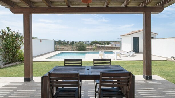 A covered patio with a pool, wooden table, and chairs.