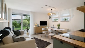 A modern living room with a sofa, a dining area with chairs, a kitchen with a counter, and a TV.