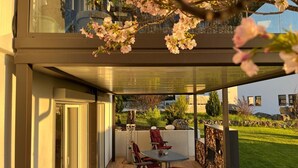 A modern outdoor patio with a glass enclosure, a dining table with chairs, and a view of a garden and building in the background.