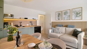 A modern kitchen with a microwave, stove, and sink. There is a dining area with a table set for two, a sofa, and a wall-mounted TV.