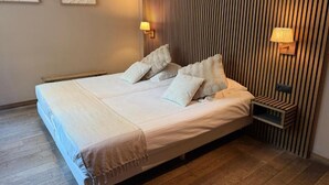 A neatly made bed with pillows, a bedside table with a lamp, and a wall with vertical lines. - Giorgi Hôtel Restaurant (Bastogne)