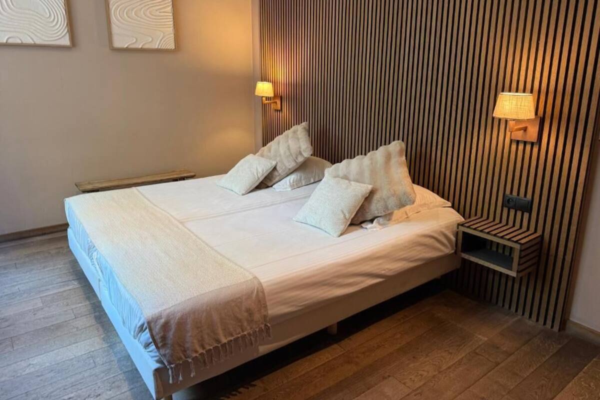 Una cama bien hecha con almohadas, una mesita de noche con lámpara y una pared con líneas verticales.