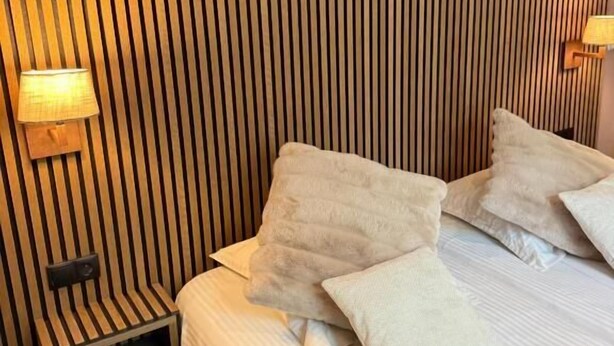 A room with a bed, pillows, a nightstand, a lamp, and a wall with vertical wooden slats. - Giorgi Hôtel Restaurant (Bastogne)