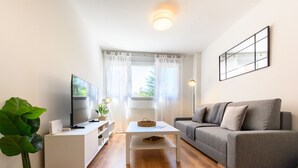 Ett vardagsrum med en platt-TV, en grå soffa, ett vitt soffbord och en stor grön växt.