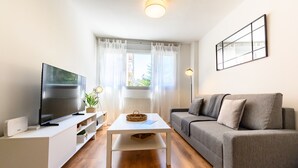Ett modernt vardagsrum med en platt-TV, ett vitt soffbord, en grå soffa och ett fönster med tunna, genomskinliga gardiner.