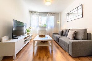 Ein modernes Wohnzimmer mit Flachbildfernseher, einem weißen Couchtisch, einem grauen Sofa und einem Fenster mit leichtem Vorhang.