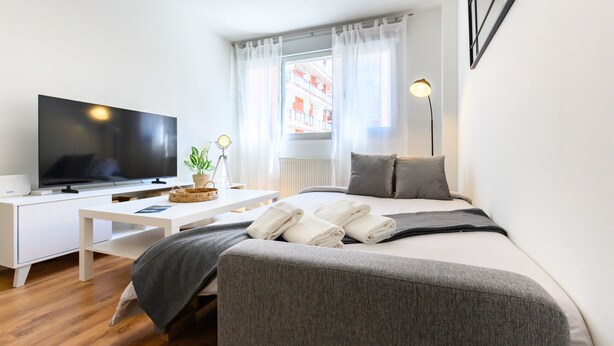 Ett modernt hotellrum med en platt-TV, ett vitt skrivbord, en säng med grå sängkläder och utsikt över byggnader genom tunna, genomskinliga gardiner.