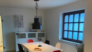 Ein Essbereich mit einem weißen Tisch, roten Stühlen, einer schwarzen Pendelleuchte und einem Fenster mit Jalousien.