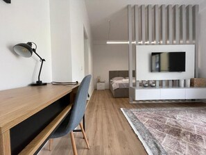 A modern living room with a wooden desk, a grey chair, a television mounted on a white wall, and a patterned rug. - Comfort Center Studio Apartments (Tirana)