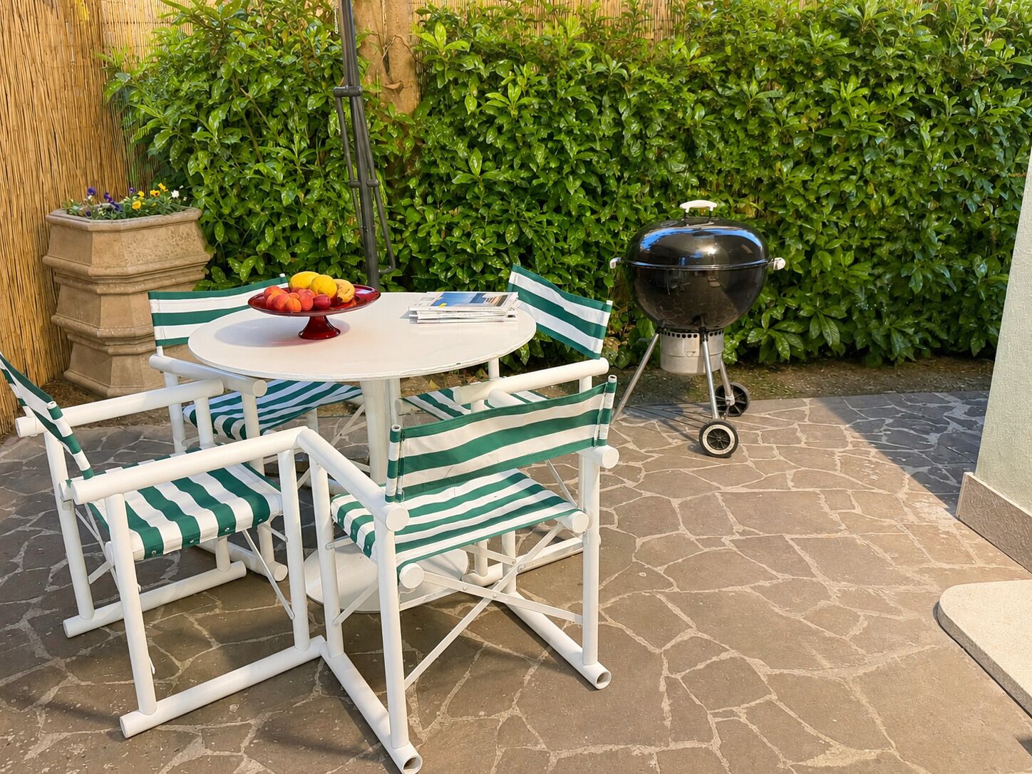 A patio with a round table, striped chairs, a fruit bowl, a grill, and a hedge in the background.