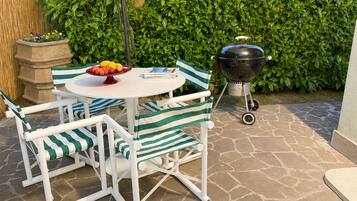 A patio with a round table, striped chairs, a fruit bowl, a grill, and a hedge in the background.
