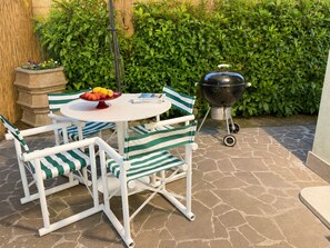 Une terrasse avec une table ronde, des chaises à rayures, un panier de fruits, un barbecue et une haie en arrière-plan.