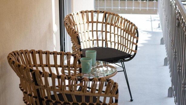 Varanda com cadeiras de vime, mesa de vidro, guarda-corpo de metal e vista para o verde.