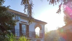 Un edificio de dos pisos con un balcón, rodeado de un jardín con diversas plantas y un camino de grava.