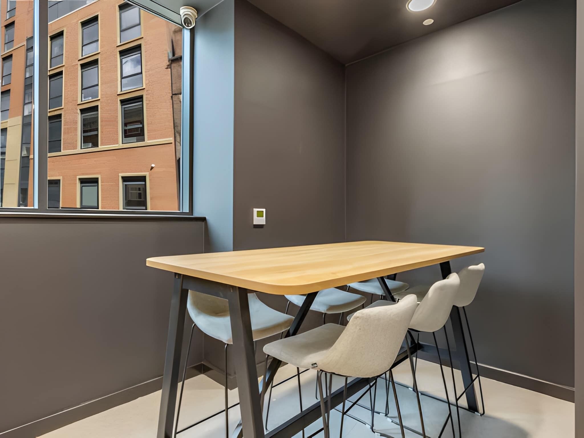 Una habitación moderna con una mesa de madera y dos sillas, una ventana con vista a un edificio y un termostato en la pared.