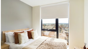 A modern bedroom with a large bed, a wooden headboard, and a view of a cityscape through a large window. - Student Only Straits Aire (Leeds)