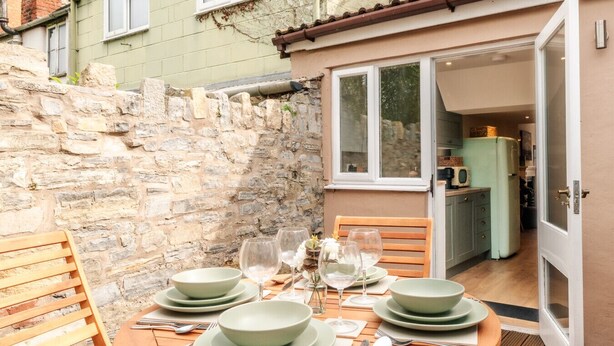 A dining table set with plates, bowls, wine glasses, and cutlery, with a stone wall and a door leading to a kitchen. - Beautiful One Bedroom Home in Glastonbury (Glastonbury)