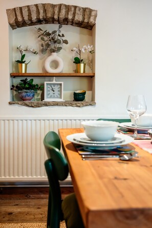 A dining area with a wooden table set for two, a green chair, a rustic brick shelf with plants and a clock, and a radiator below.