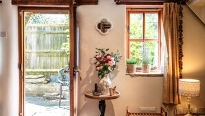 A cozy room with a wooden door, a small table with a vase of flowers, a window with curtains, and a lamp. - Beautiful Two Bedroom Cottage in Oxfordshire (Warborough)