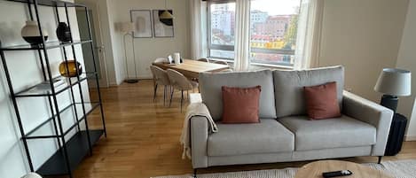 Una sala de estar moderna con un sofá gris, una mesa de centro, una estantería y un comedor con una mesa y sillas de madera.
